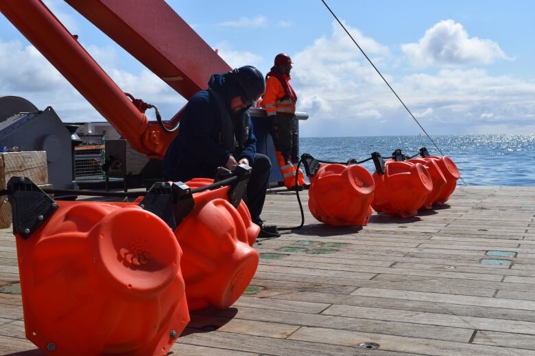 A researcher is kneeling near several bright orange buoys, the atlantic ocean visible in the background.