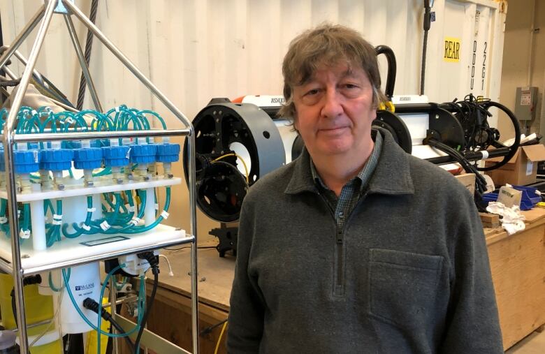 An oceanography professor is shown standing in a lab with equipment behind him.