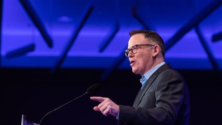 A man with short hair and glasses addresses a crowd off-camera.