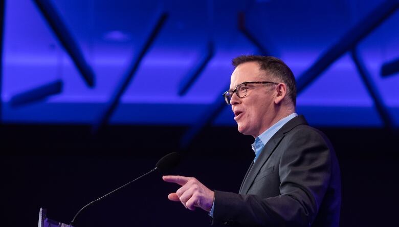 A man with short hair and glasses addresses a crowd off-camera.