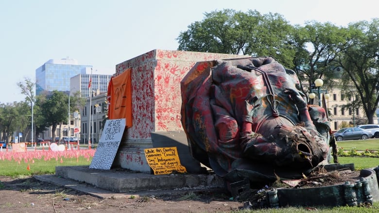 A statue of a sitting woman, with its head removed, lies on its back next to the pedestal it had been standing on. The pedestal and statue are covered in red handprints. 