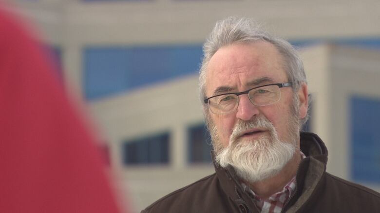 Man with short grey hair, white beard, and glasses stands outside the beige and blue NL power building. He has a serious look on his face.