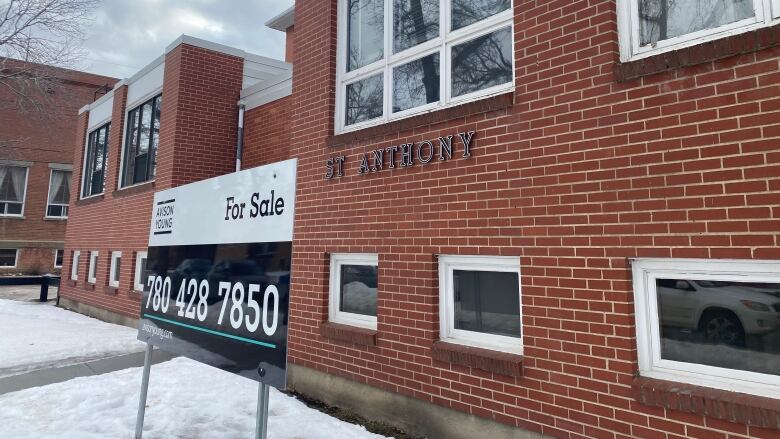 A for sale sign stands in front of a brick school building. There is snow on the ground in front of the building.