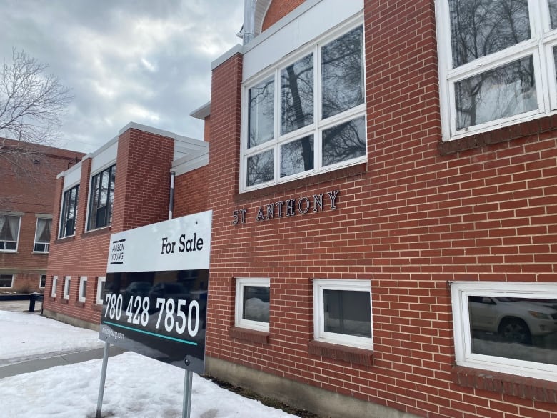 A for sale sign stands in front of a brick school building. There is snow on the ground in front of the building.