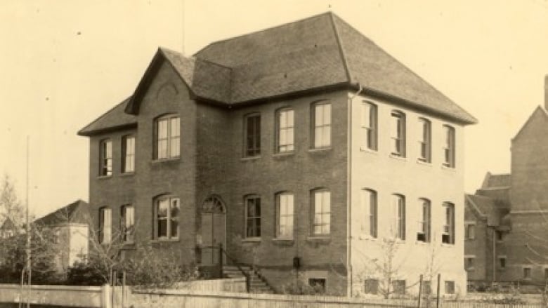 A very old black and white photo of a small square brick school building with a shingled roof.