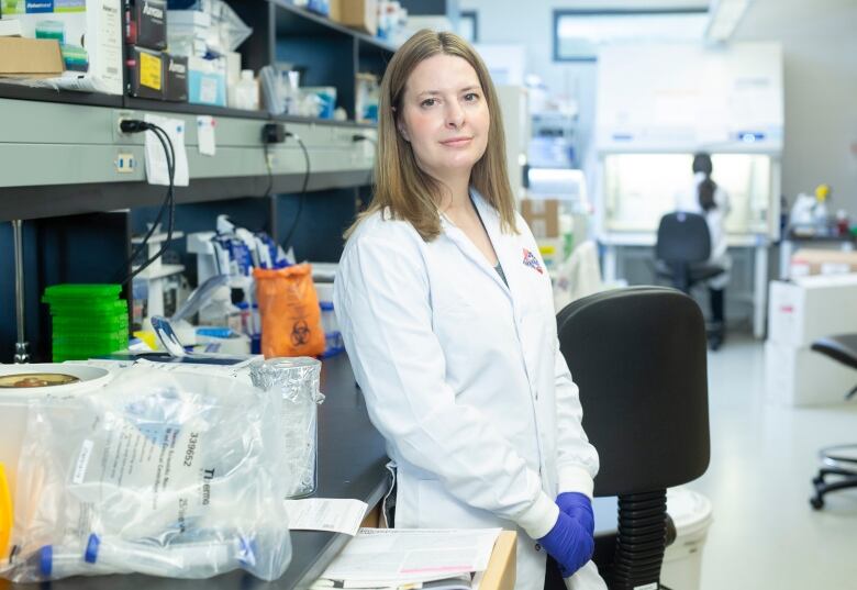 A woman in a lab coat leans against a counter with lab equipment in the background