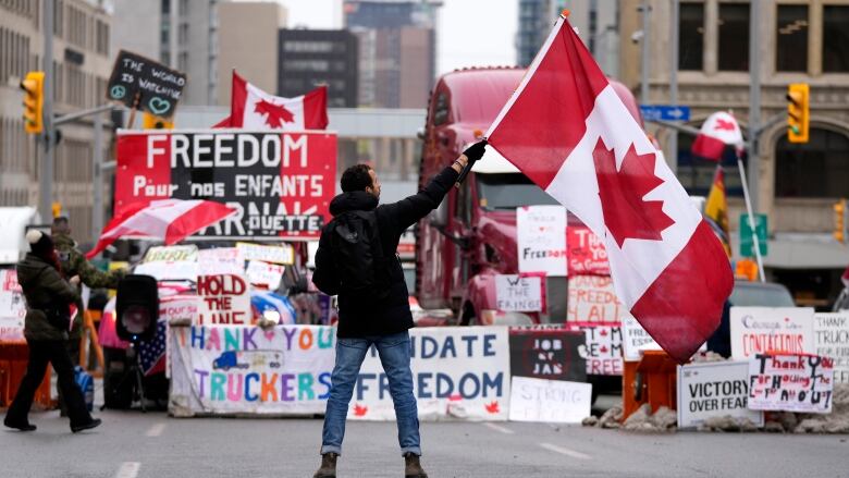 A man stands in the middle of the street waving a Canadian flag, facing a line of large trucks decked out in hand-drawn signs, including a colourful banner that reads 