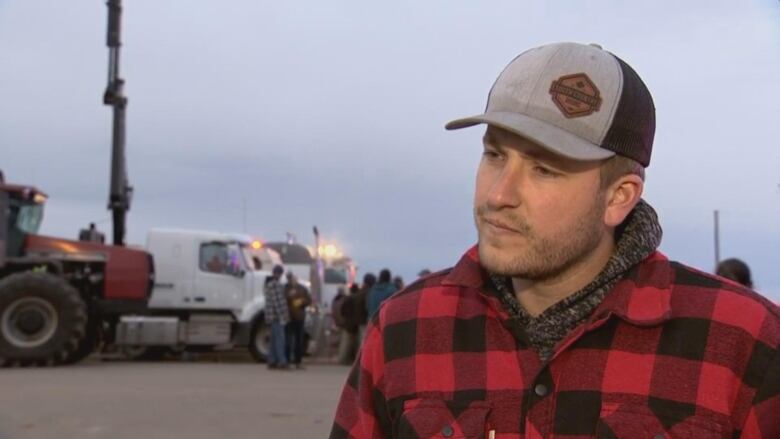 A while man in a red and black buffalo check jacket with a ball cap on. He's standing in front of a tractor and a truck.