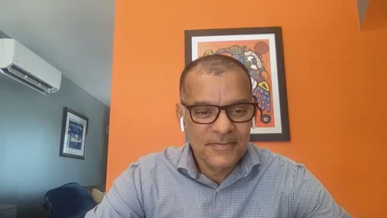 A Black man wearing glasses sits in front of an orange wall