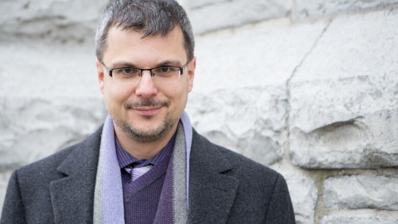 A man in black-framed glasses in a purple collared shirt and dark grey jacket.