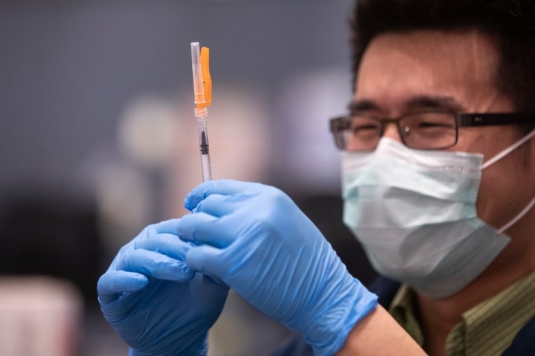 A man with a mask and blue gloves holds a syringe in his hand.