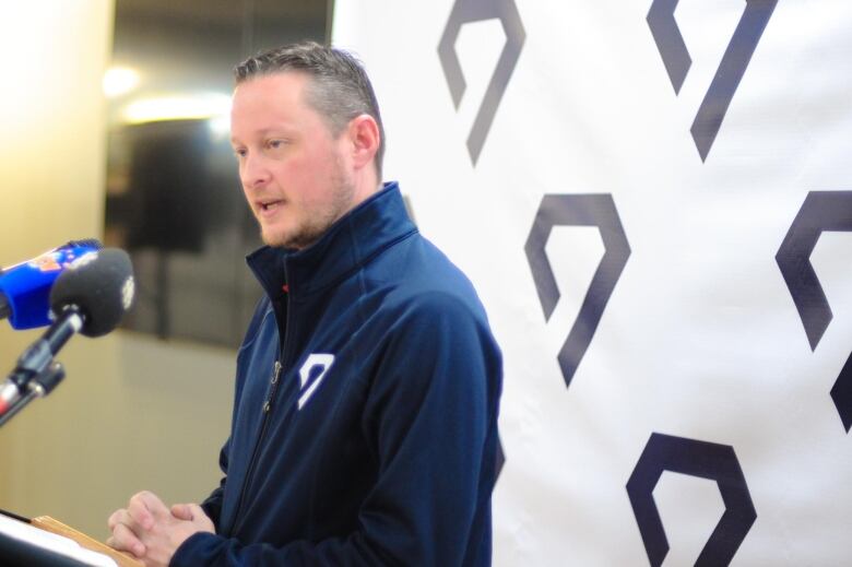 A man in a blue jacket speaks into a microphone while facing to the left with a logo in the background.