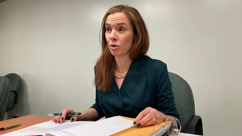 A woman sits behind a table with an open binder in front of her.