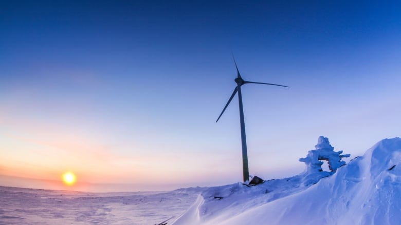 A wind turbine stands tall in in the middle of an Arctic tundra, with an inuksuk in the foreground.