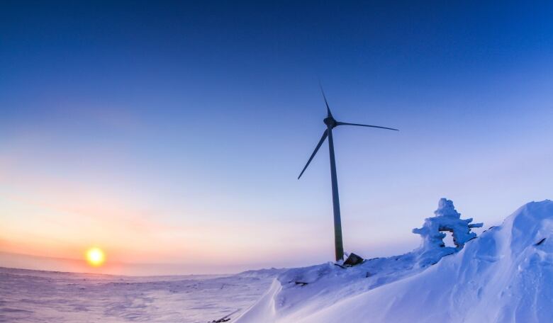 A wind turbine stands tall in in the middle of an Arctic tundra, with an inuksuk in the foreground.