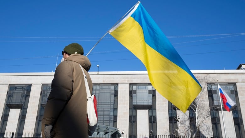 A man is seen from behind holding a Ukrainian flag in front of a building where the Russian flag is flying.