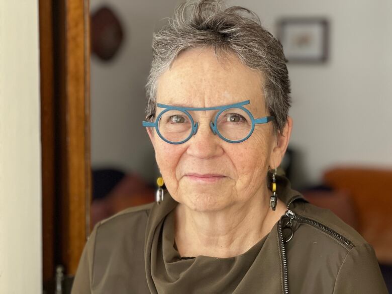 A woman with short hair and turquoise-coloured glasses smiles while looking at the camera.