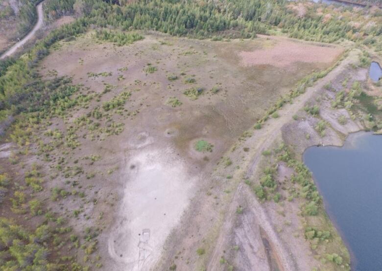 An aerial photo shows a barren piece of land bordered by trees on three sides and a body of water.