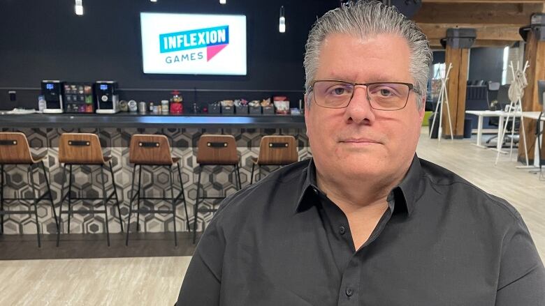 Scott Nye in black button-up shirt looks at camera. Behind him is a snack bar with a hanging television displaying the logo for Inflexion Games.