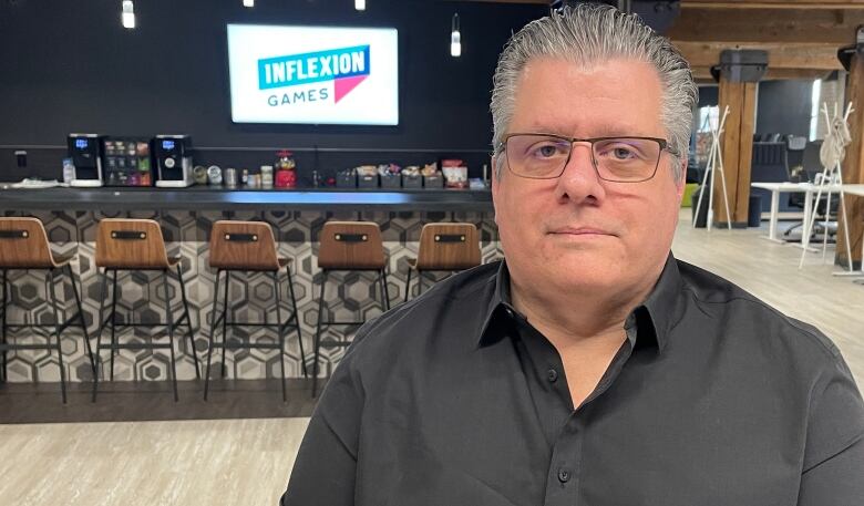 Scott Nye in black button-up shirt looks at camera. Behind him is a snack bar with a hanging television displaying the logo for Inflexion Games.