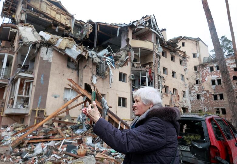 A woman with grey hair holds a cellphone in front of herself. Apartment buildings half ripped apart are shown in the background.