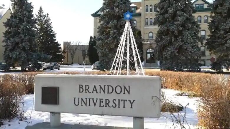 A concrete sign that reads 'Brandon University' is seen surrounded by snow and plants.