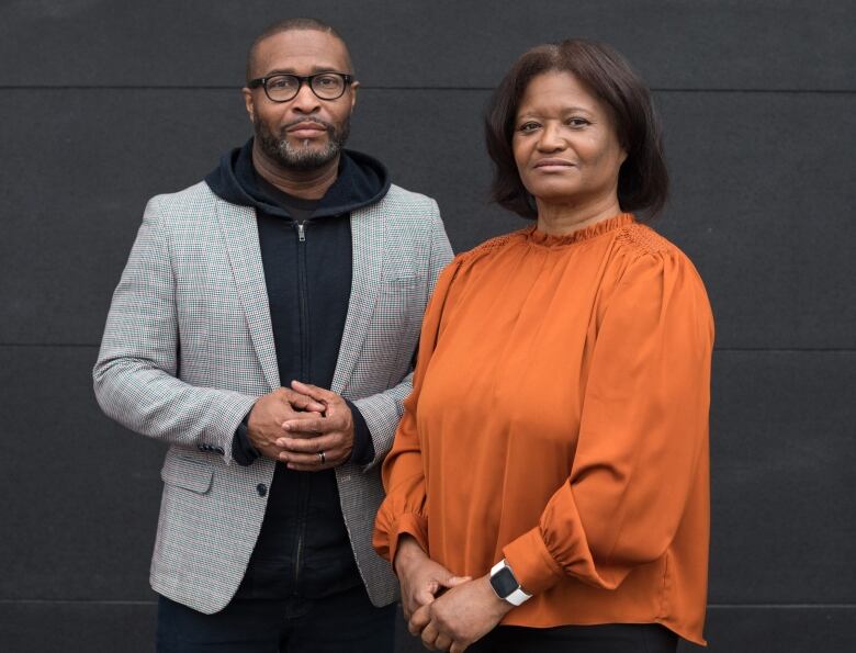 A Black man and woman standing beside each other. The man is wearing glasses and a grey blazer over a black hoodie. The woman is wearing and orange blouse.