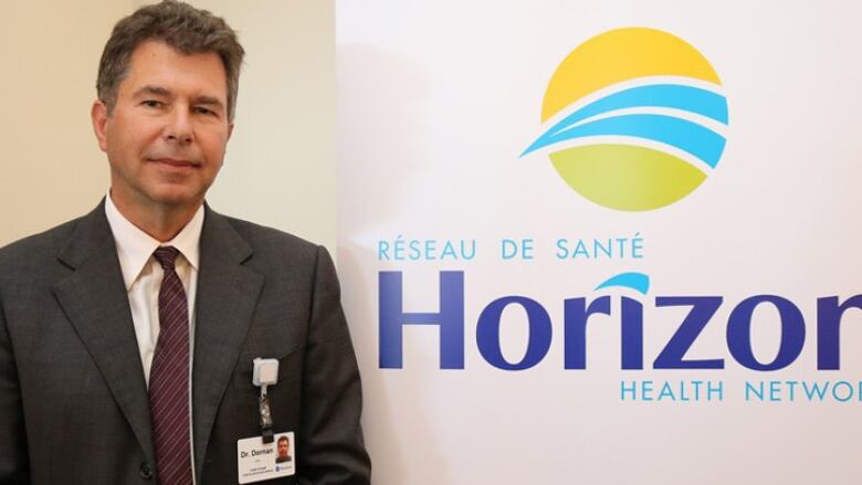 A man wearing a suit and an ID badge on his pocket poses in front of a wall with a large logo for Horizon Health Network. 