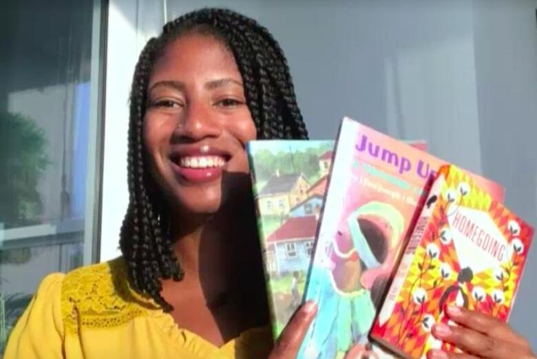 A Black woman with braids is smiling and holding up three books