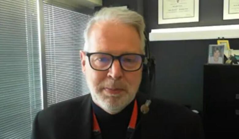 Dr. Daniel Gregson wears black glasses, a black suit, and  a red lanyard as he looks at the camera. Degrees are hanging on the grey wall behind him.