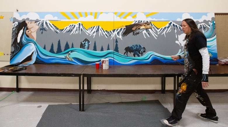 Man walks in front of a mural that shows a flowing river, mountains and a sunny sky. The river appears as though it is flowing from a woman. 