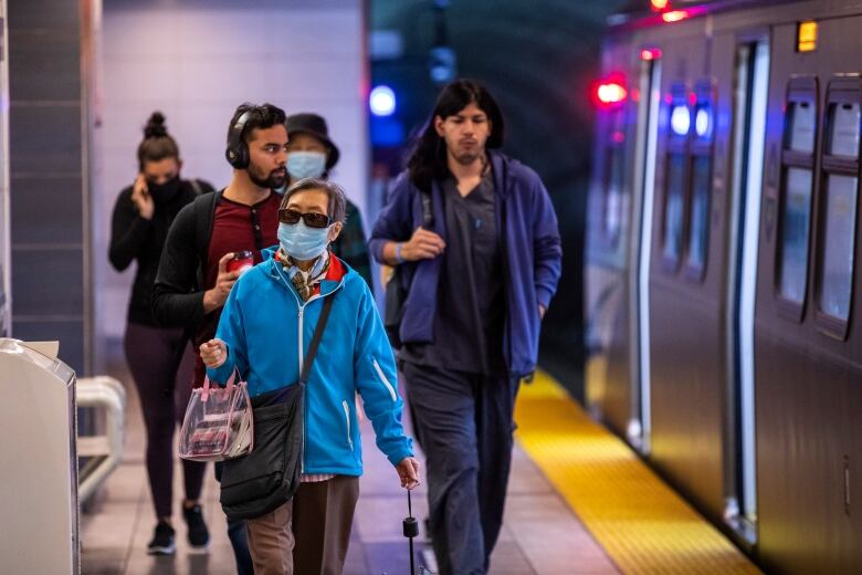 A group of people walk alongside a train in a subway. Some of them are wearing facemasks and others are not.