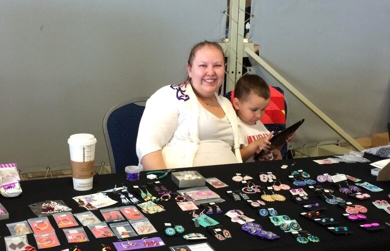 An indigenous woman in a white shirt and sweater sits in front of a table of colourful beaded crafts and smiles as a young boy plays with a computer tablet.