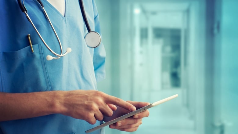 A closeup shows a doctor in a hospital hallway holding a tablet. 