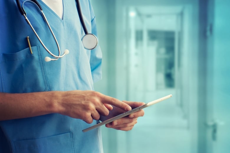 A closeup shows a doctor in a hospital hallway holding a tablet. 