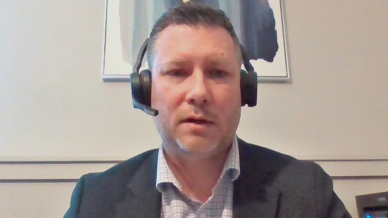 A Zoom screenshot shows a man in a suit jacket and dress shirt wearing a headset looking into the camera. He's seated in a room with beige walls and there's a piece of art behind him that's various shades of blue and grey.