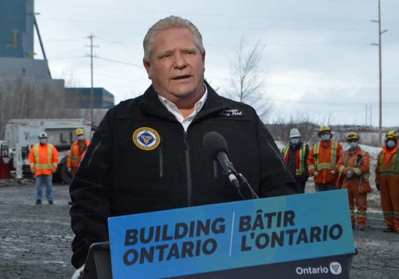 A man stands at a podium that says, Building Ontario, with workers in orange coveralls in the background.