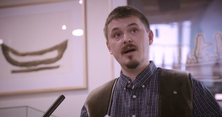 A man stands before a microphone with a white wall and art in the background. 