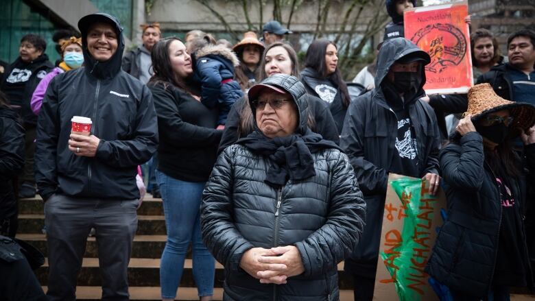 A group of people, many of whom are Indigenous, stand on steps. Some of them are carrying colourful signs.