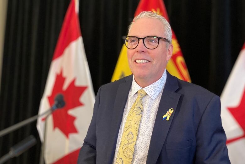 A man smiling in front of a New Brunswick flag with two Canadian flags on either side of it