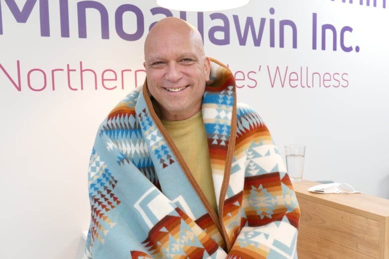 A man with a shaved head smiles, wrapped in a colourful patterned blanket.