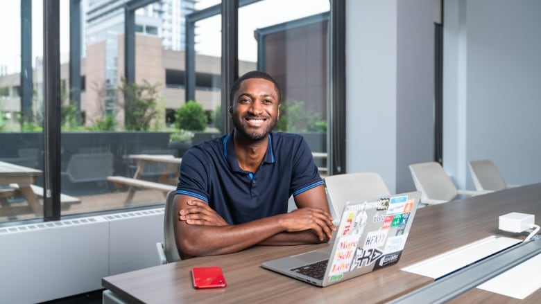 A man in a dark blue short-sleeve shirt smiles while sitting at a desk in front of a laptop.