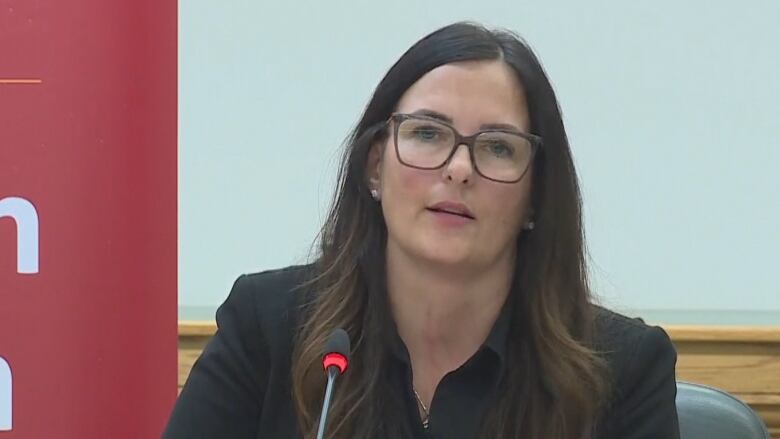 A white woman with long brown hair is wearing glasses and an all-black suit. She is sitting in front of a microphone.