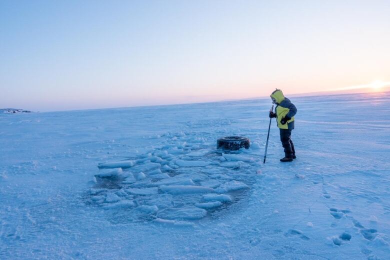 A person standing on sea ice looks at a big, partially frozen hole. 