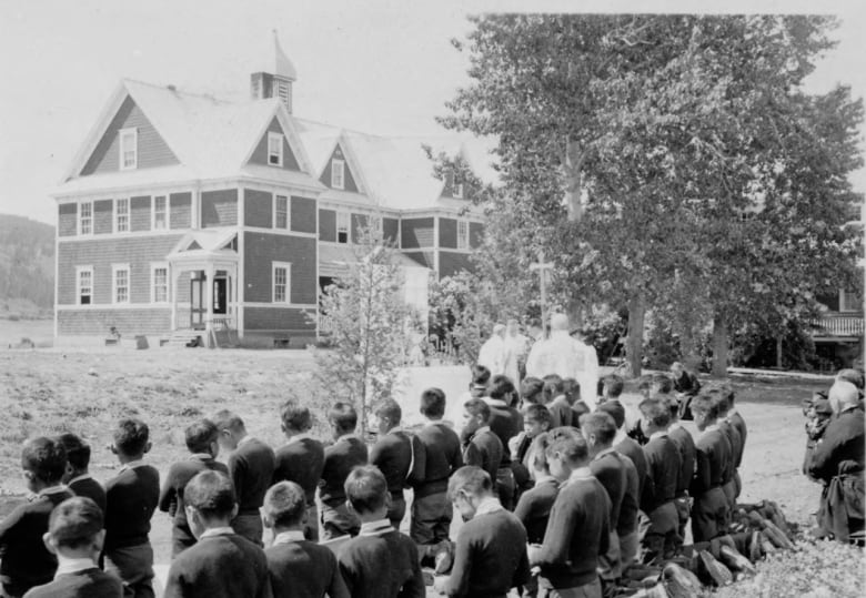 Children kneeling gathered in a group outside a schoolhouse in an undated black and white photo.