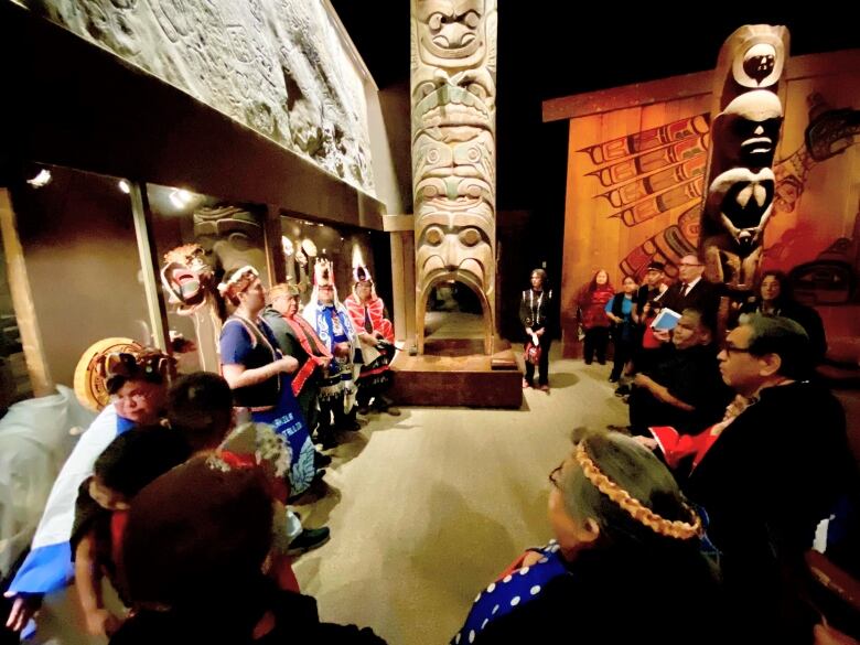 Members of the Nuxalk nation in ceremonial garb with other dignitaries present are arrayed around an intricately carved totem pole that is surrounded by native art in a museum.