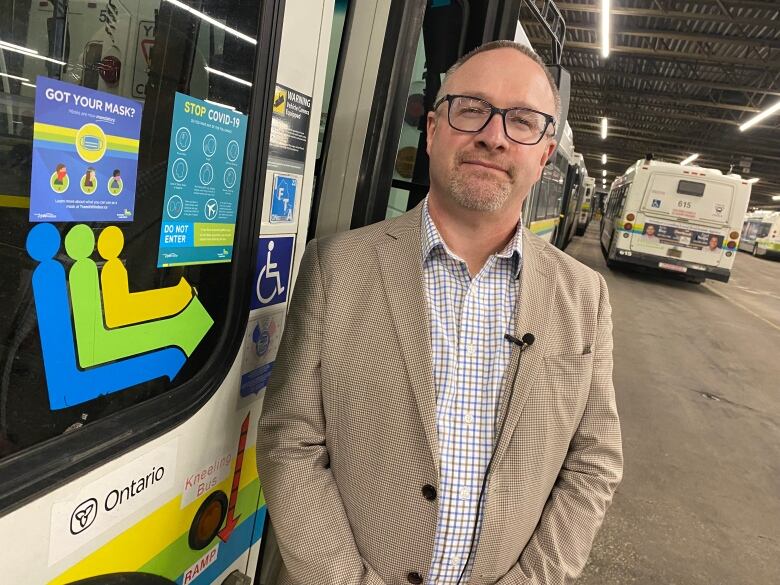 A bald man wearing glasses and a brown suit standing next to a public transit bus