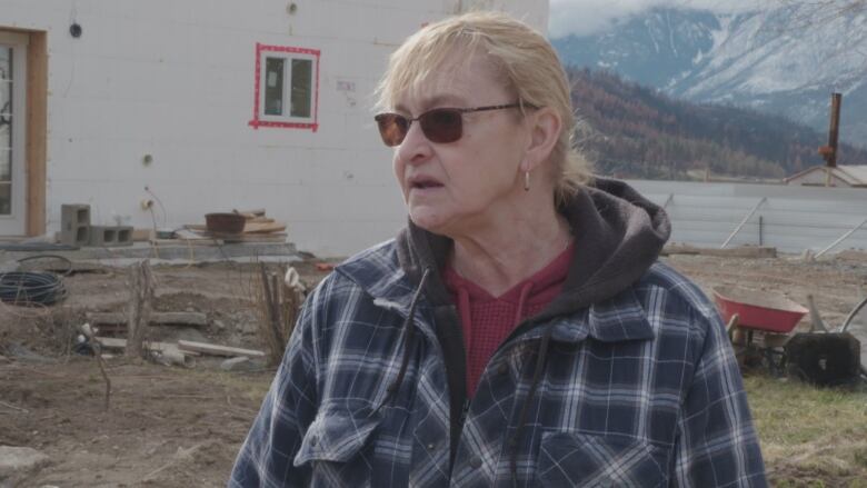 A woman in a flannel coat framed by mountains and a rundown white wooden building with wood haphazardly piled outside speaks to a reporter off-camera.