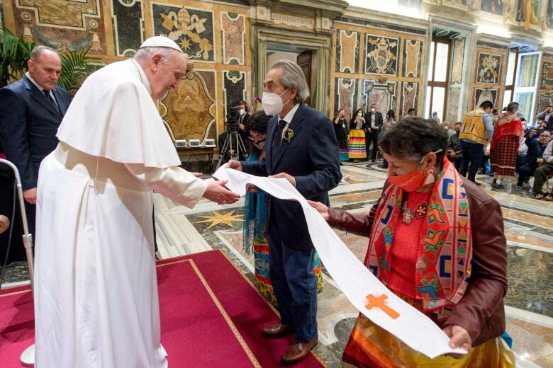 A long, white leather garment that is beaded with orange crosses is pictured being gifted to the Pope by two people.