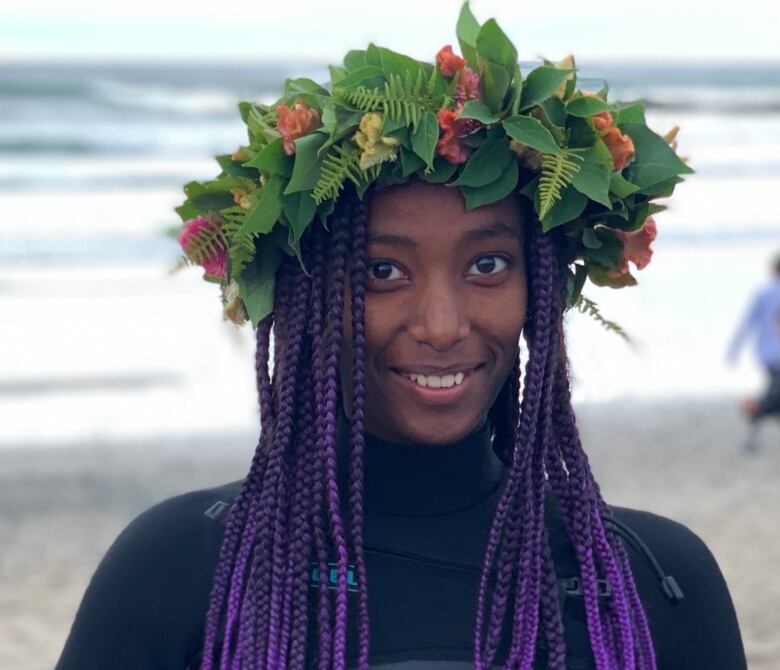 A Black woman wearing with purple braids wearing a flower crown and surfing gear